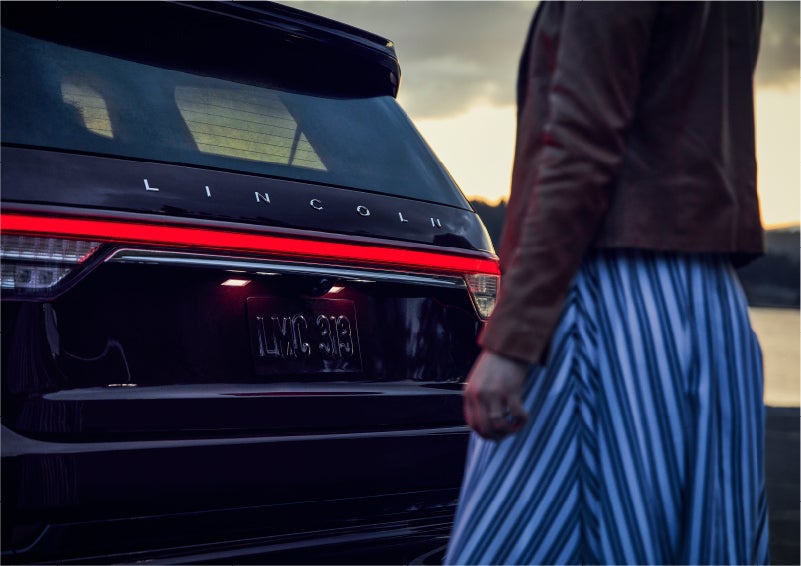 A person is shown near the rear of a 2023 Lincoln Aviator® SUV as the Lincoln Embrace illuminates the rear lights | Gary Yeomans Lincoln in Daytona Beach FL